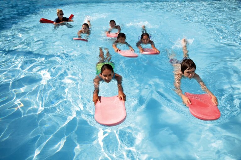 A young child sits on the edge of an indoor pool smiling, with a swim instructor offering gentle encouragement nearby.