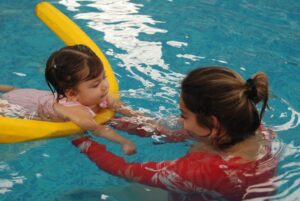 A swim instructor guiding a young child through a gentle water activity in an indoor pool.