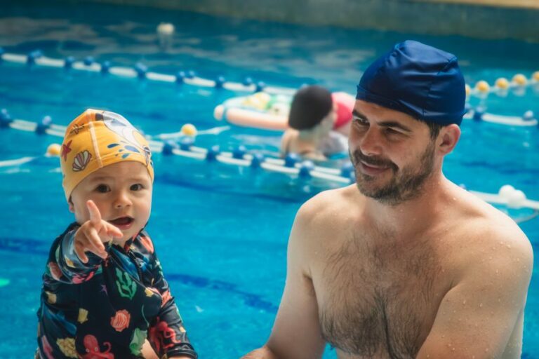 Child practices blowing bubbles in shallow water while a swim instructor provides calm support.